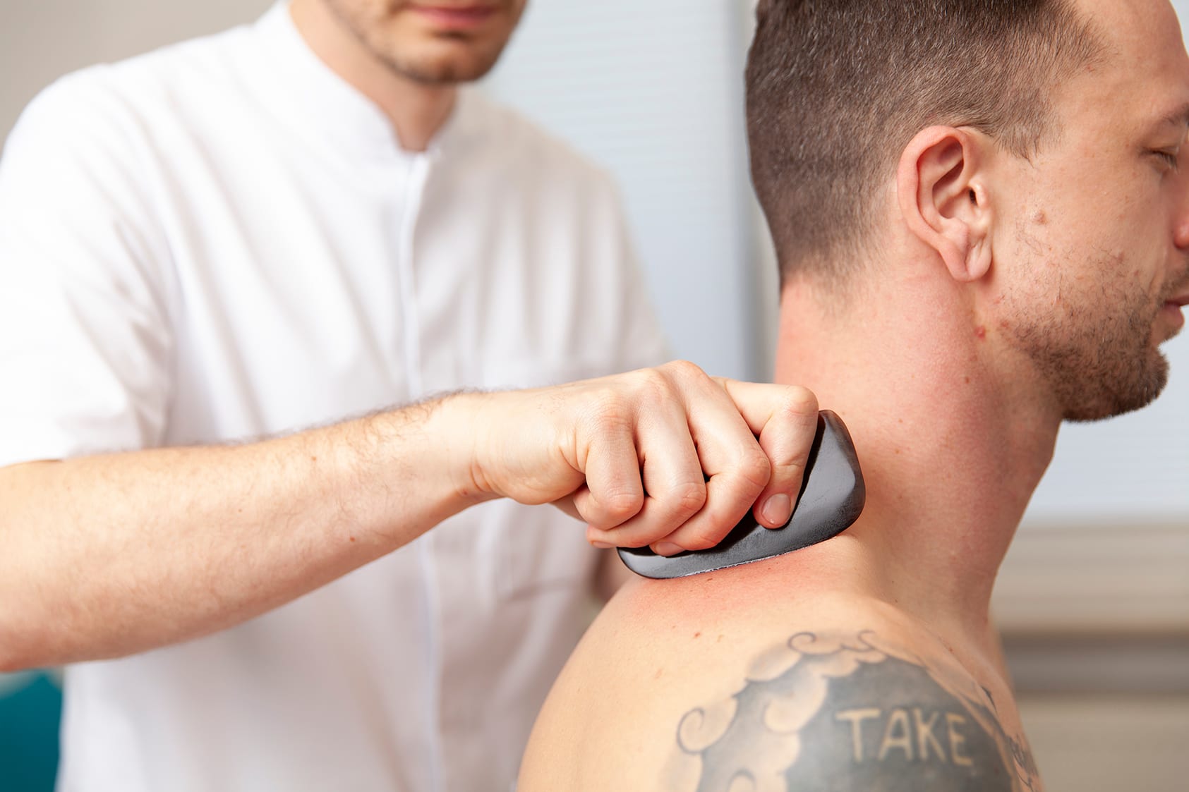 Heilpraktiker Rick Putzer führt eine Gua-Sha-Behandlung am Nacken eines Patienten durch.