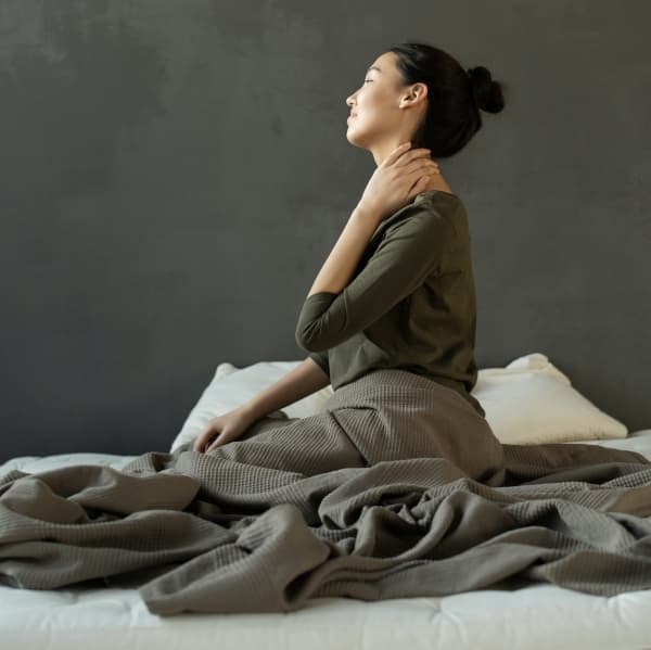 Eine Frau sitzt im Bett und fasst sich an den verspannten Nacken.